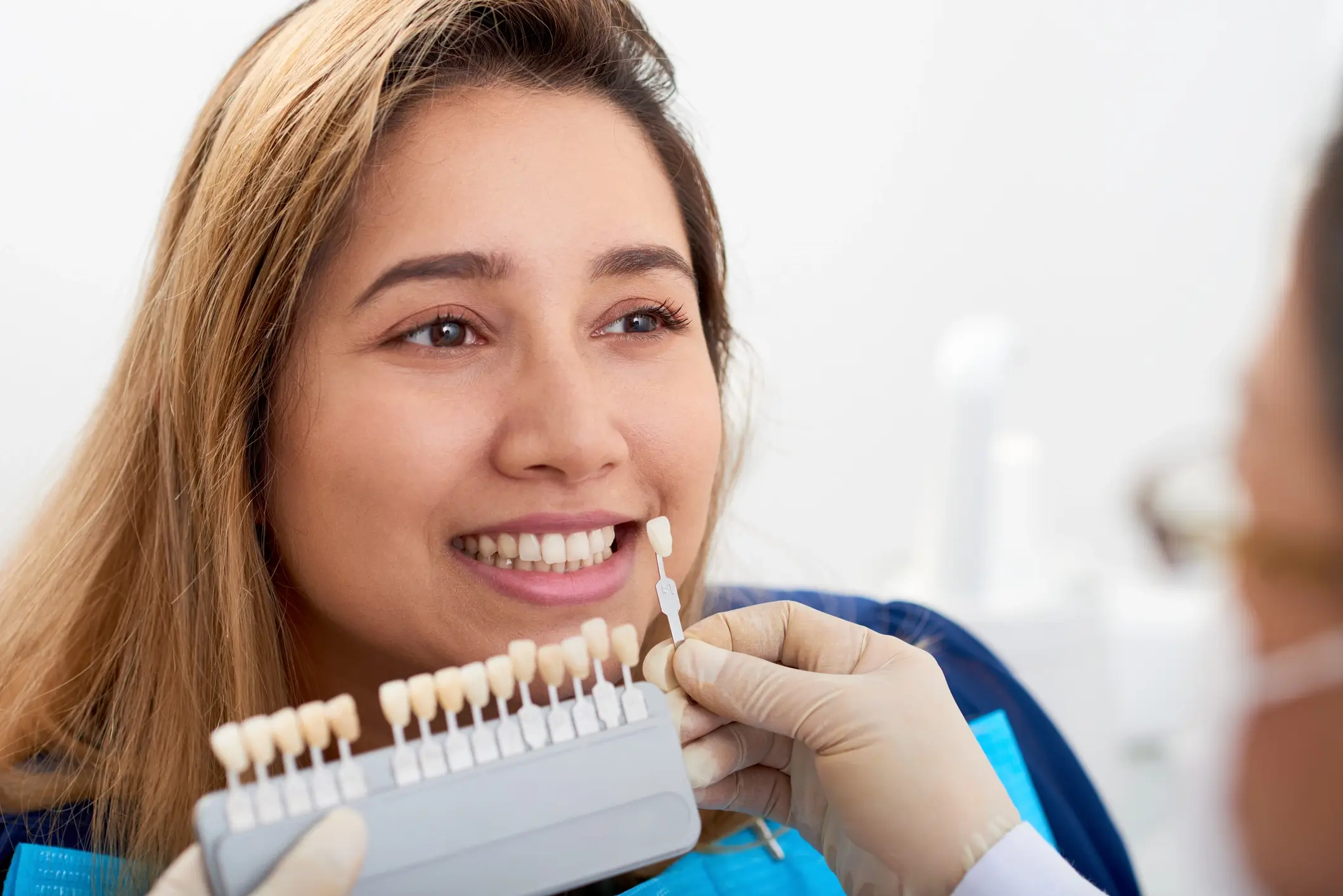 Cosmetic Dentistry Veneer Woman smiling while a dentist holds a shade guide to match teeth color for cosmetic veneers, in a dental office setting.
