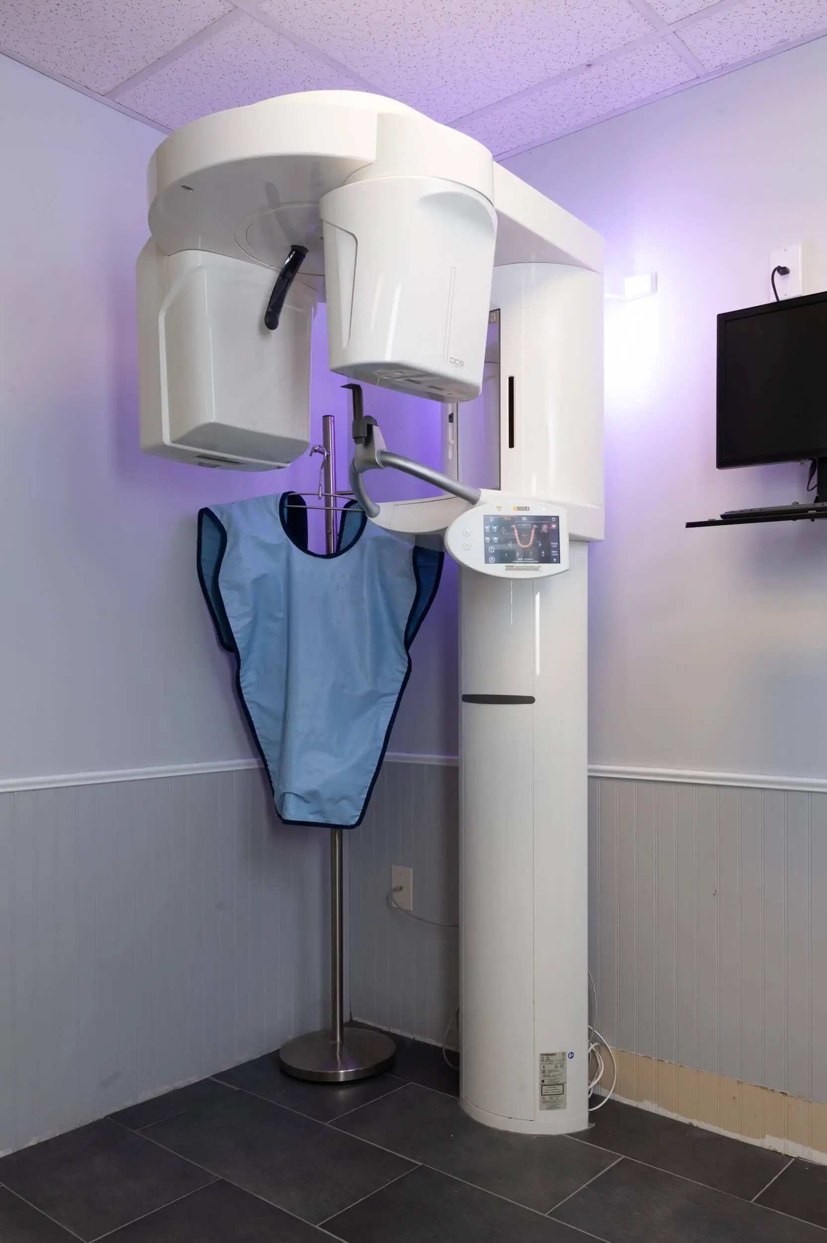 Cone beam 3D imaging machine in dental office, featuring a blue protective apron and digital control panel, highlighting advanced imaging technology for precise dental diagnostics and treatment planning.